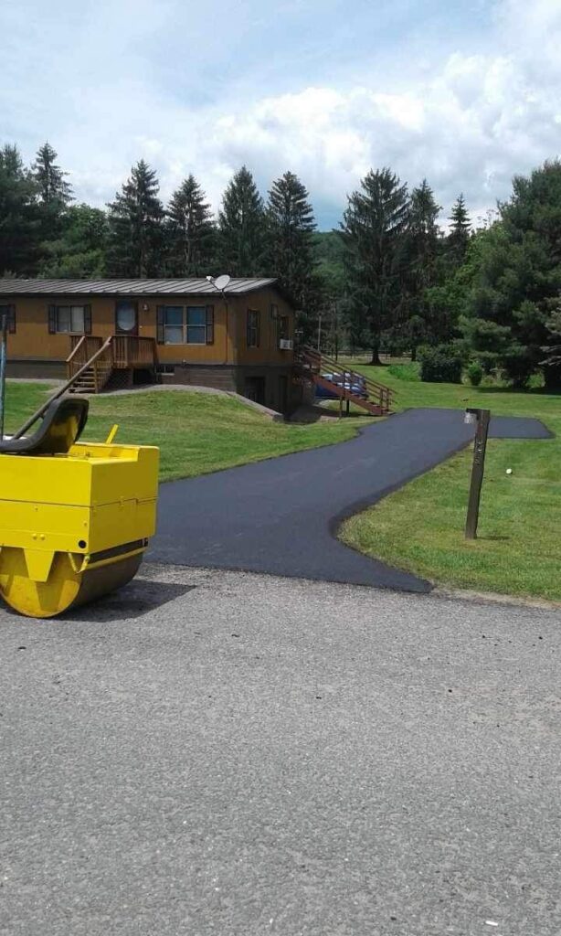 A freshly paved driveway leading to a house with a front porch, surrounded by a well-maintained lawn. A yellow road roller is visible in the foreground.