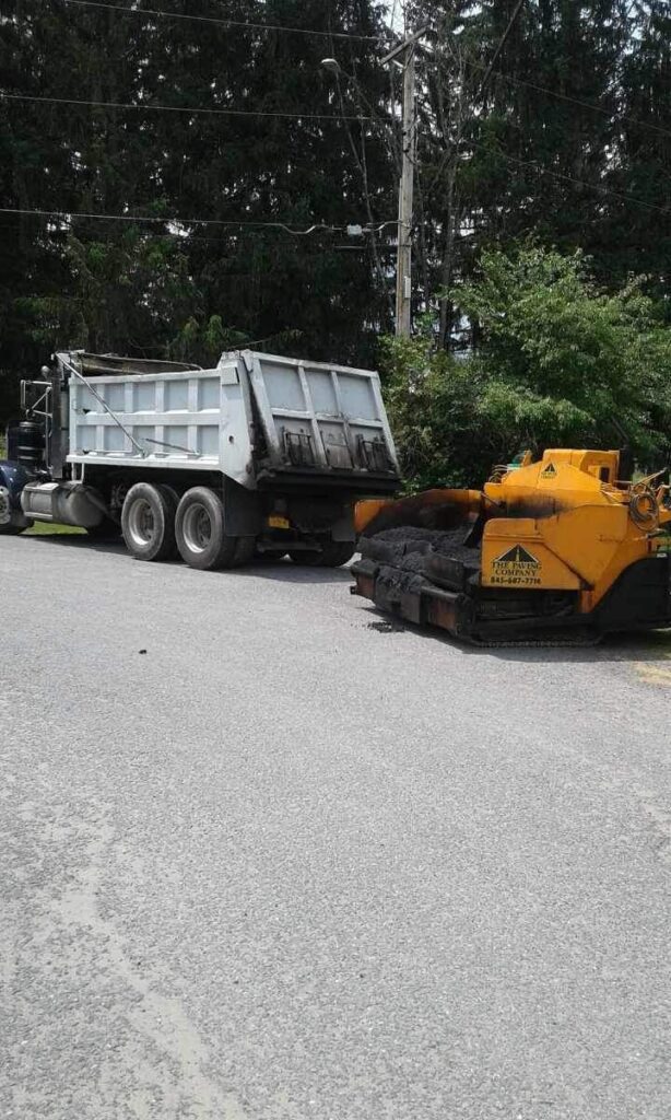A dump truck is parked beside an industrial machine labeled "Paver". Both are located on a paved road near a vegetated area.