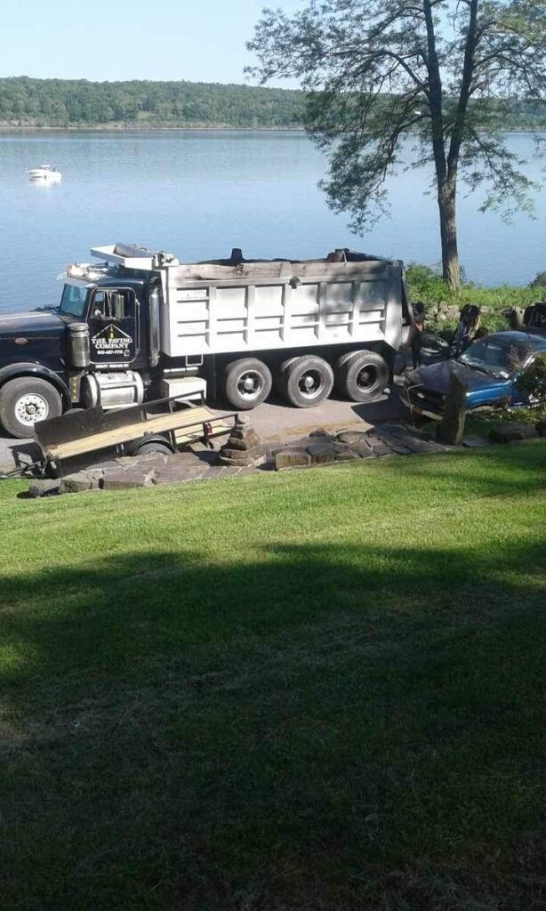 A dump truck and a parked car are on a paved area near a lake with a large tree. A boat is visible on the lake in the background.