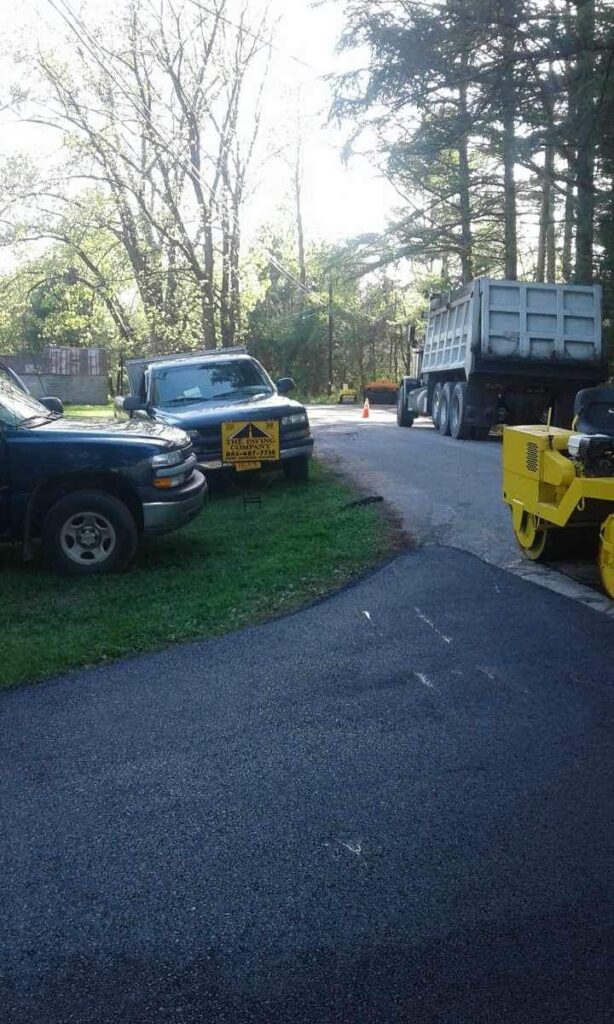 Two trucks parked on the left side of a road with a construction sign between them, and a dump truck and a roller on the right side of the road near trees and vegetation.