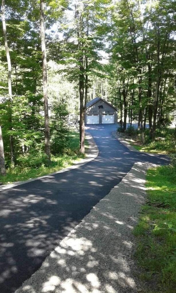 A paved driveway curves through a sunlit forest, leading to a two-car garage at the end, surrounded by tall trees.