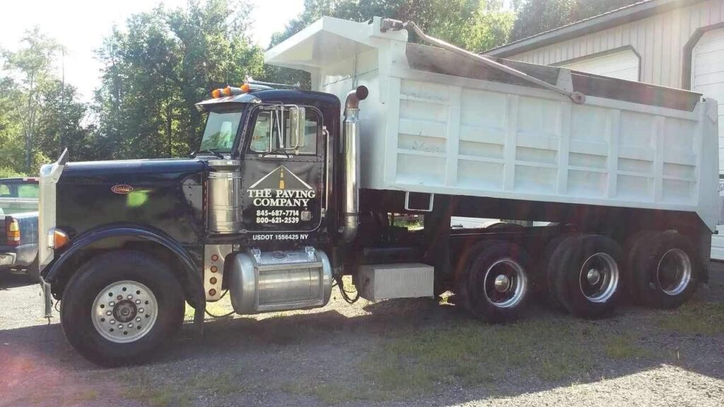 A black and white dump truck with "The Paving Company" logo and contact details parked beside a building.