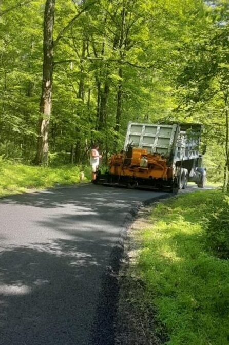 A worker operates a vehicle laying new asphalt on a forest road, with trees and greenery surrounding the scene.